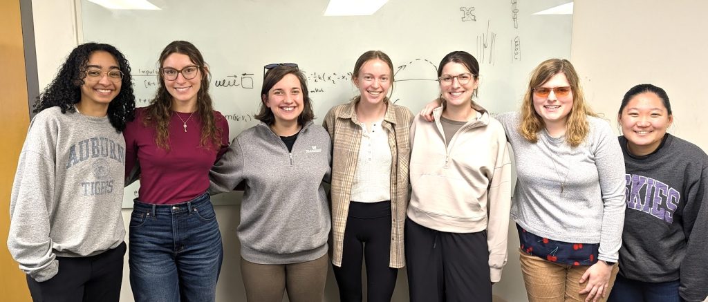 A group of seven people stand side by side in a classroom or lab space, posing for a photo in front of a large whiteboard. The individuals are standing close together with arms around one another, and they are dressed in casual clothing.