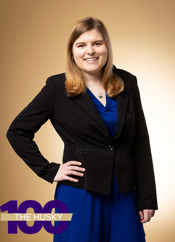 Studio portrait shows Mia Hoffman standing in a black blazer and blue dress against a gold gradient background; the Husky 100 logo is visible at the bottom of the image.
