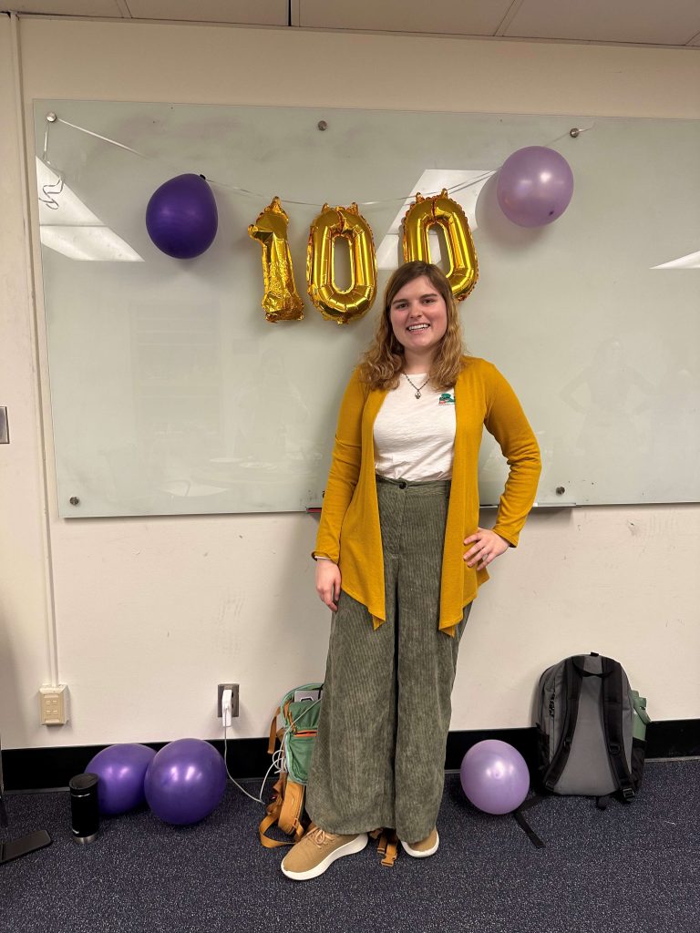 Person stands in a classroom in front of gold “100” balloons and purple balloons, posing for a photo to celebrate Husky 100 recognition.