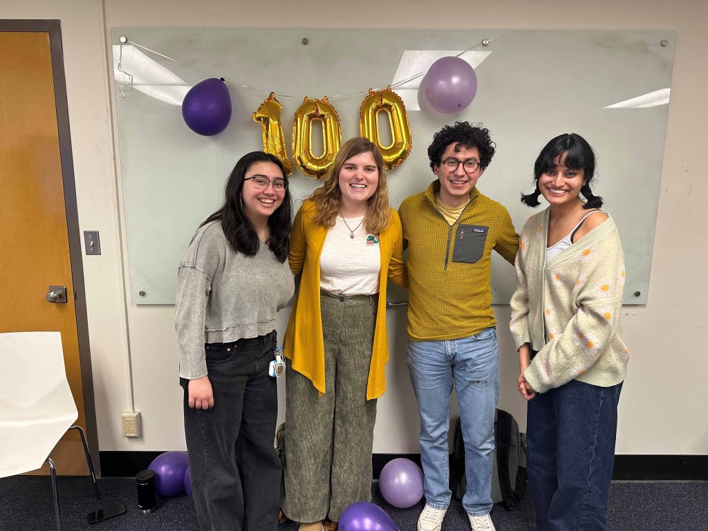 Five people stand together in a conference room, smiling for a group photo in front of gold “100” balloons and purple balloons, celebrating Husky 100 recognition.