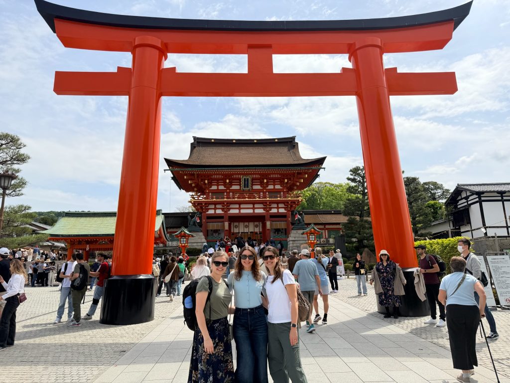 Three people pose in front of a large red torii gate at a busy shrine in Japan, with a traditional temple building and visitors in the background.
