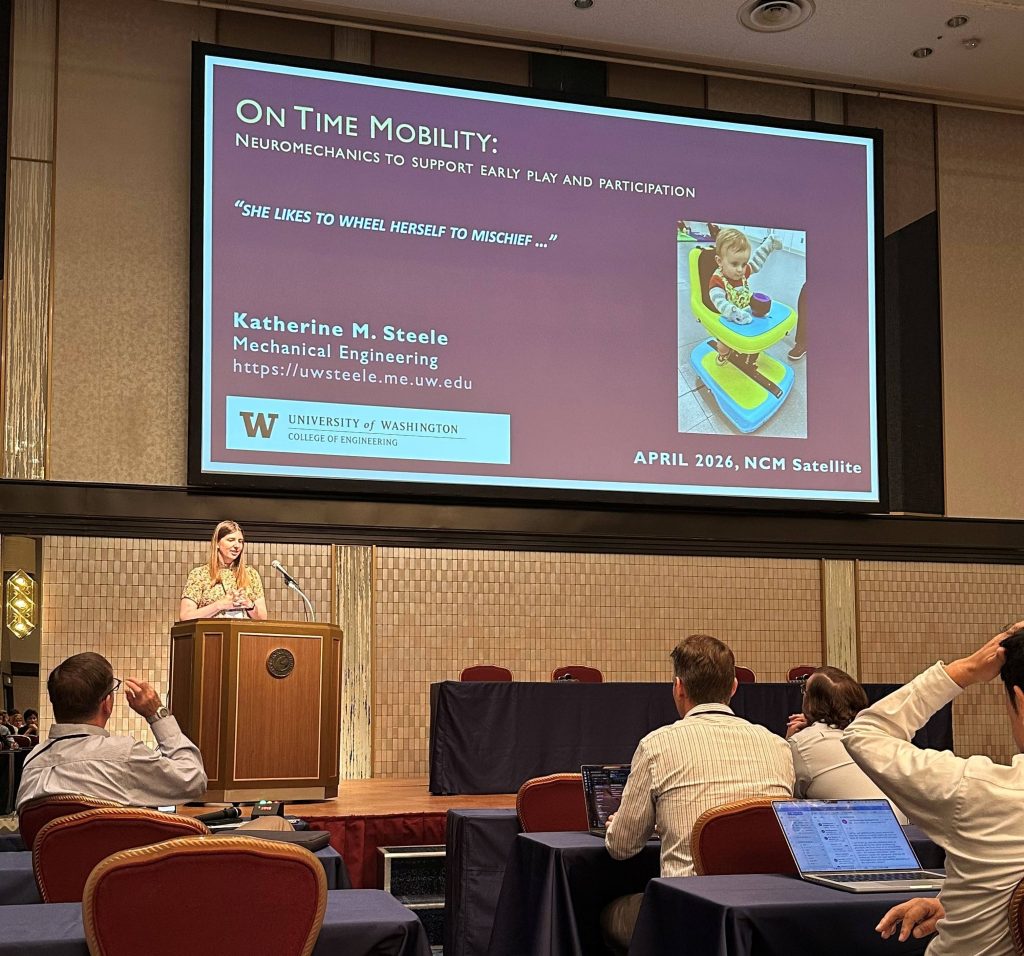 Dr. Kat Steele stands at a podium presenting to an audience, with a large slide reading “On Time Mobility: Neuromechanics to support early play and participation” displayed on a screen at an NCM conference session.