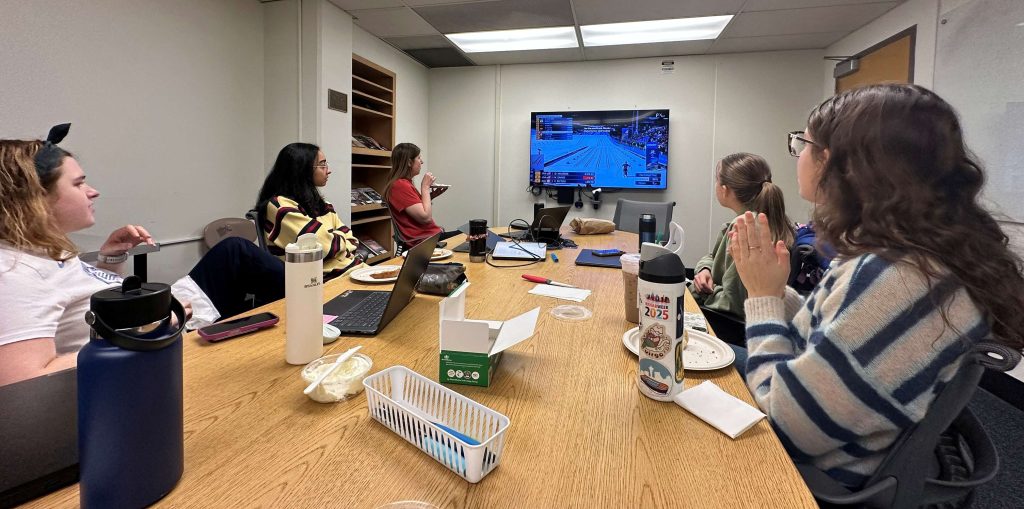 Group gathers in a conference room to watch a Para Nordic ski race on a television during a watch party.