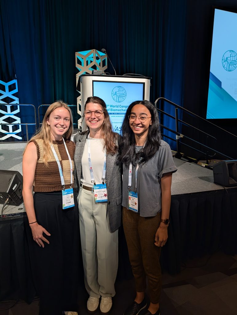 Ally, Katie, and Mackenzie stand together after giving podium presentations. Ally stands to the left, and is wearing a brown sweater and has blonde hair. Katie stands in the middle, wearing a white shirt with grey suit jacket and has brown hair. Mackenzie stands to the right wearing a grey t-shirt and dark brown hair.