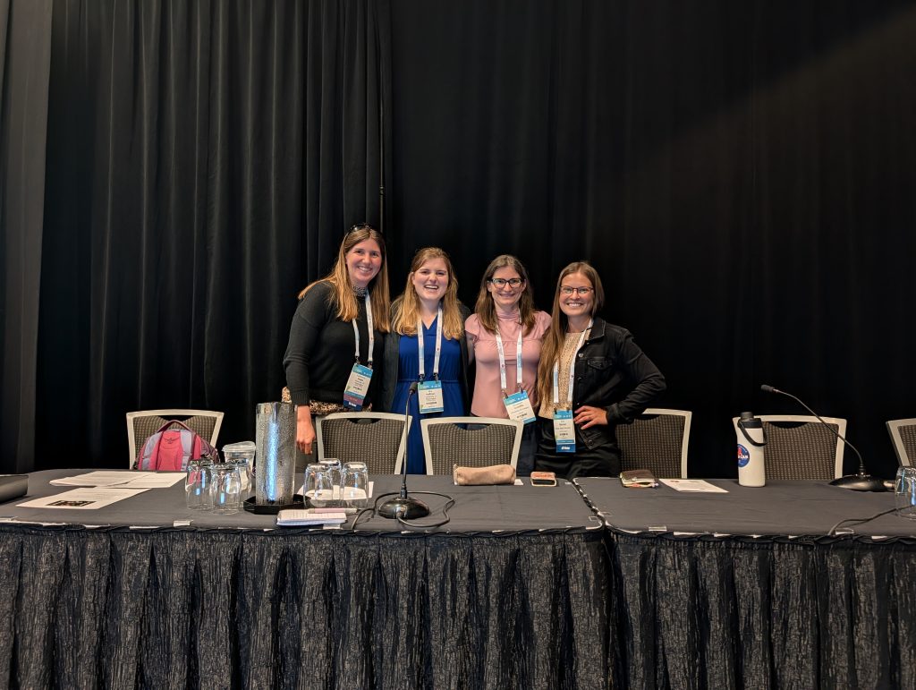 Kat and Mia stand with Elizabeth Condliffe and Erin Mannen. All women are wearing professional clothing and standing behind the presenter's table.