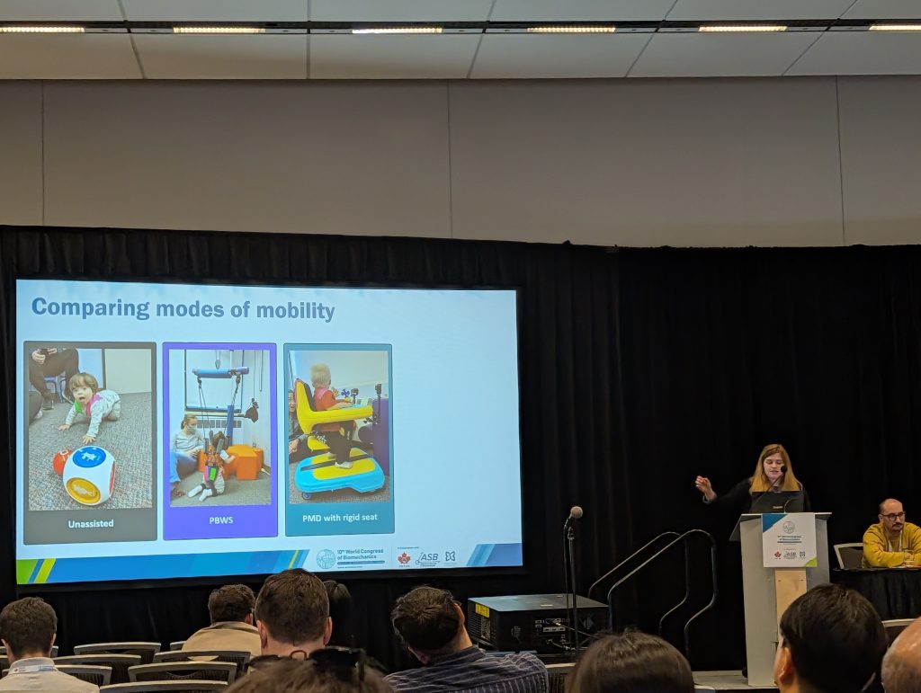 Mia stands to the right of a powerpoint slide during her presentation. The powerpoint slide depicts young children using mobility devices