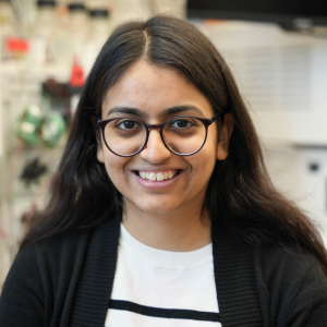 Prisha has dark brown hair and is wearing glasses, a white striped shirt, and black sweater.