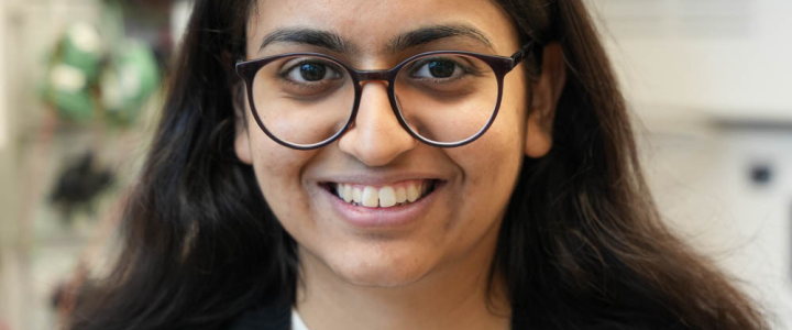 Prisha has dark brown hair and is wearing glasses, a white striped shirt, and black sweater.
